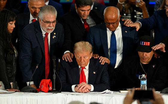 Then-Republican presidential nominee Donald Trump sits with his head bowed and is surrounded by Latino leaders also with bowed heads.