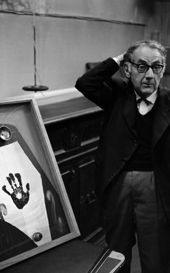 Artist Man Ray — photographed in his Paris studio — stands next to one of his works, titled simply “Self-portrait.” The work is a 1963 remake of an original from 1916 that was lost or destroyed, but considered one of the world’s first Dada objects — even before the term was coined. Ray described his piece as, “the butt of much joking. On a background of black and aluminum paint I attached two electric bells and a real push button. In the middle, I had simply put my hand on the palette and transferred the paint imprint as a signature. Everyone who pushed the button was disappointed that the bell did not ring.”  