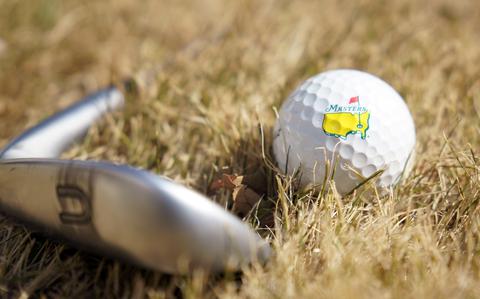 A close-up view of a golf ball with the yellow Masters logo and a golf club sitting on grass.