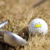 A close-up view of a golf ball with the yellow Masters logo and a golf club sitting on grass.