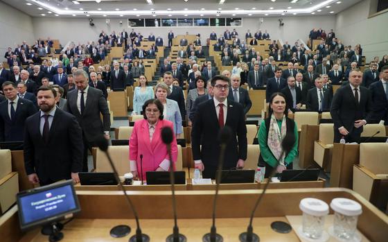 Russian members of Parliament stand at attention to observe the national anthem, with a speaker’s podium in the photo foreground.