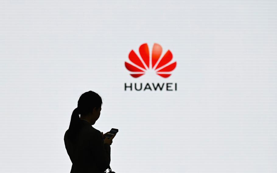 The silhouette of a person looking at their phone while in front of a screen or white poster, with Huawei’s logo seen just right-of-center.