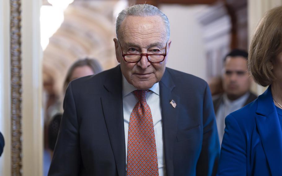 Chuck Schumer, wearing a dark blue suit and red tie, walks out of a meeting room in the U.S. Capitol building.