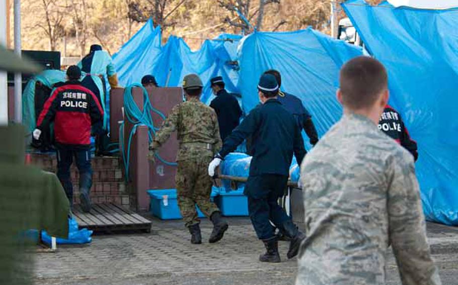 An airman looks on as morgue workers, police officers and Japan Self-Defense Force troops bring a body to a morge. Soldiers from Logistics Task Force 35, along with a few airmen from the 374th Civil Engineering Squadron from Yokota Air Base, established two shower stations near the Ono Civic Center shelter in Higashimatsushima, Japan, which also borders a morgue.