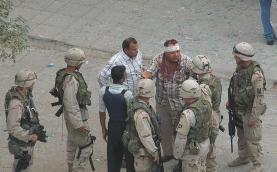 A wounded Iraqi civilian, with a bandaged head and bloody shirt, talks to American soldiers near the scene of a car bombing outside the Baghdad Hotel. Within a half hour of the blast American troops and Iraqi police had set up a temporary perimeter about a block from the scene, later reinforcing it with concertina wire.