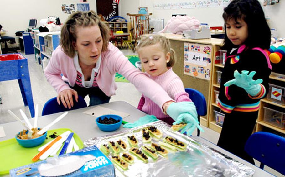 ure Start Program teaching assistant LaVenna McCune helps pupil (and daughter) Ashlin McCune place a piece of “ants on a log” on a platter as Matea Herrera looks on Tuesday at Sasebo Elementary School. Ants on a log are celery sections, stuffed with peanut butter and topped with raisins (the ants).
