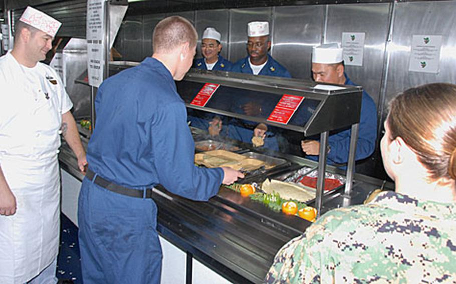 Petty Officer 2nd Class Dennis Adams, left, a culinary specialist on the USS Harpers Ferry, looks on as sailors and Marines get dinner Saturday. The ship has to double and sometimes triple the amount of food made when Marines are onboard.