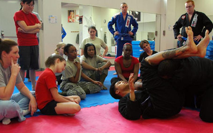 Students, including Kim Kincaid and her daughter Claire, both seated to the left, watch as their instructors demonstrate how to perform an arm bar from the guard position during a sexual assault prevention self defense class at Yokota Air Base.