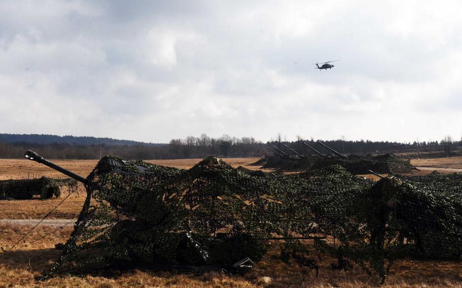 British artillery pieces prepare to fire as a U.S. Apache helicopter flies above during Exercise Dynamic Front 18 at Grafenwoehr, Germany on Wednesday, March 7, 2018.