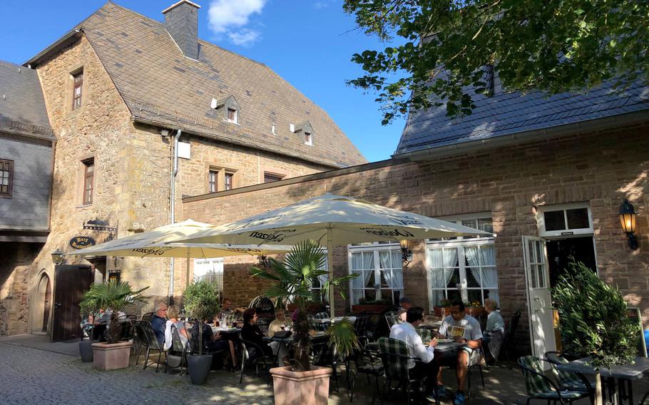 The large patio at BurgRestaurant at Lichtenberg Castle, in Thallichtenberg, Germany, is shaded by umbrellas and trees.