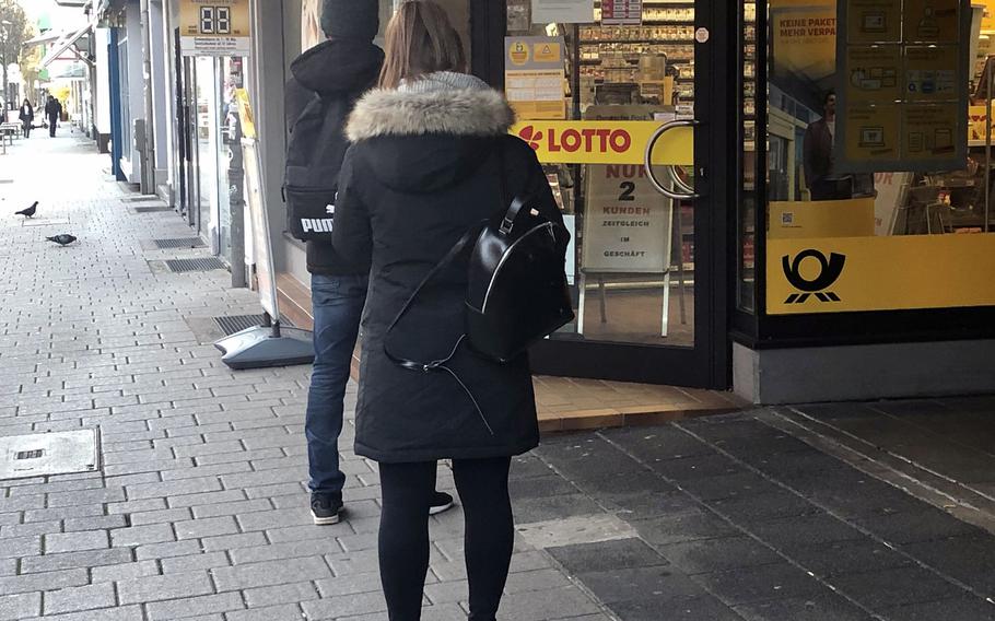 Two people stand about a meter apart as they wait to go into a shop in Kaiserslautern, Germany, on Monday, March 23, 2020. A sign inside the shop says only two customers can be inside at any given time, reflecting tougher social distancing rules announced a day earlier by Chancellor Angela Merkel.