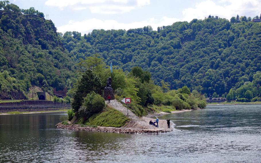 The statue of Lorelei, the siren that according to lore, lured sailors to crash their boats on the rocks at the narrowest point of the Rhine River, stands at the end of a harbor breakwater. At top left, the viewing platform of the Lorelei can be seen with its flagpoles, high above the river.