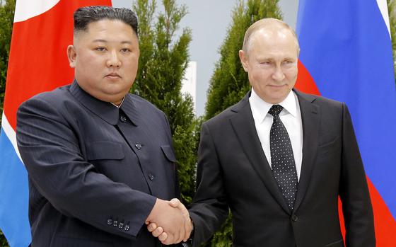 Vladimir Putin and Kim Jong Un shake hands while standing in front of Russian and North Korean flags.