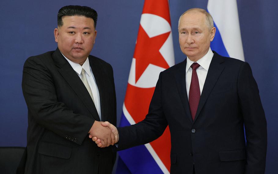 Vladimir Putin and Kim Jong Un shake hands while standing in front of Russian and North Korean flags.