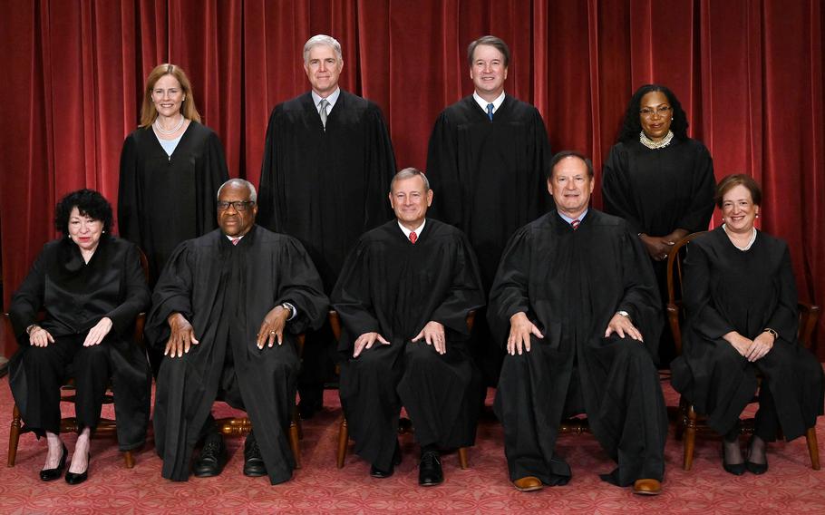 The nine justices of the U.S. Supreme Court pose in their jucidical robes, with five seated in a row and four standing behind.