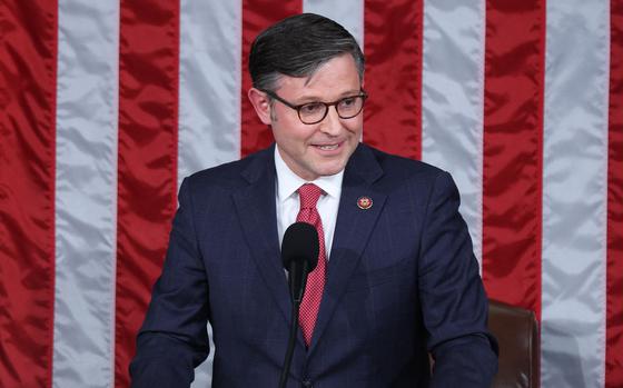 Mike Johnson, wearing a dark suit and red tie, speaks into a microphone while standing in front of a large U.S. flag.