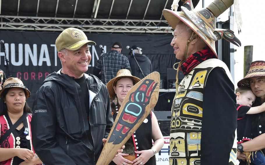 A Naval officer holds a canoe paddle with tribal painting next to an Alaskan native in traditional tribal attire.